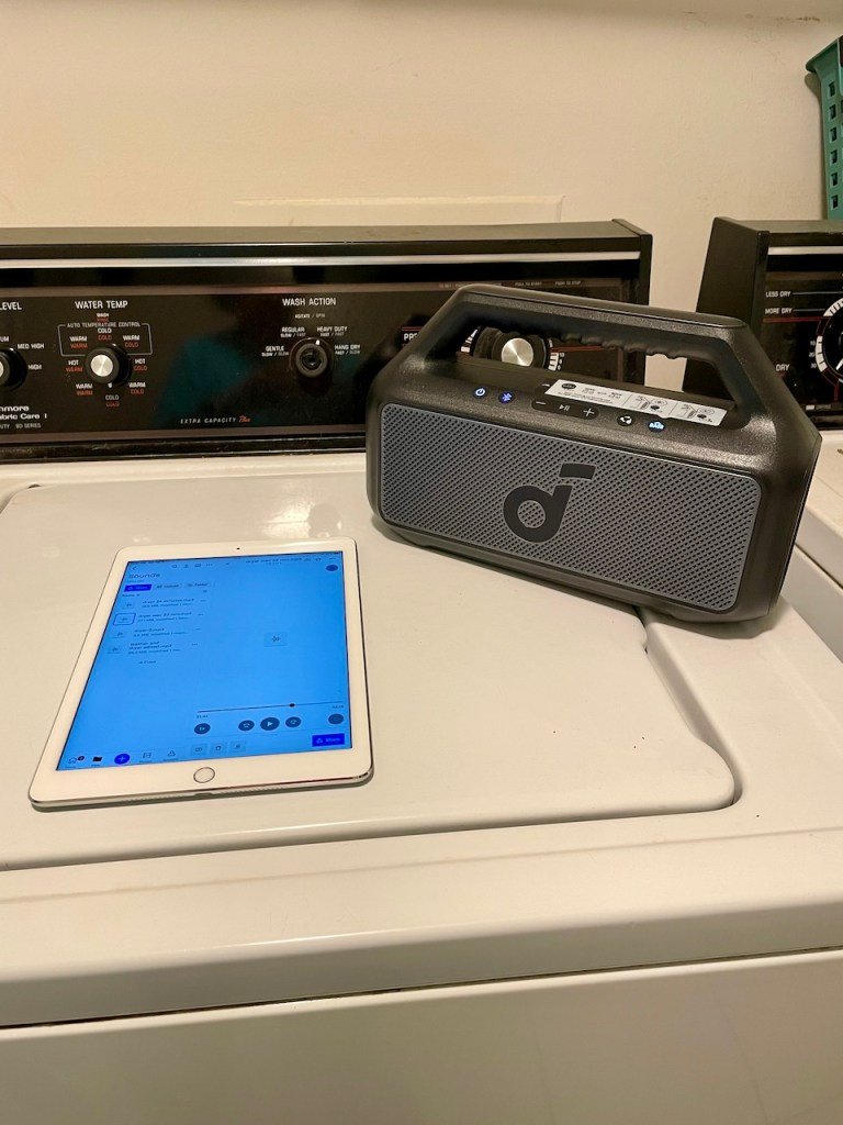 An iPad and a black bluetooth speaker with a handle on sitting together on top of a washing machine