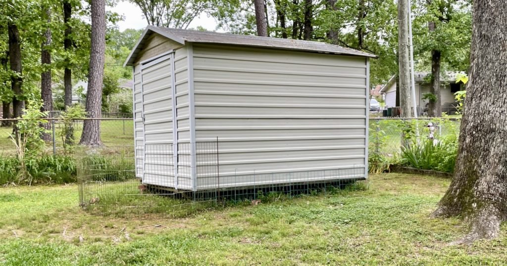 A small shed of white corrugated cardboard aluminum sits in a grassy yard. It sits on concrete blocks, resulting in rooms under the shed. There are fences that block access to the space of larger animals.